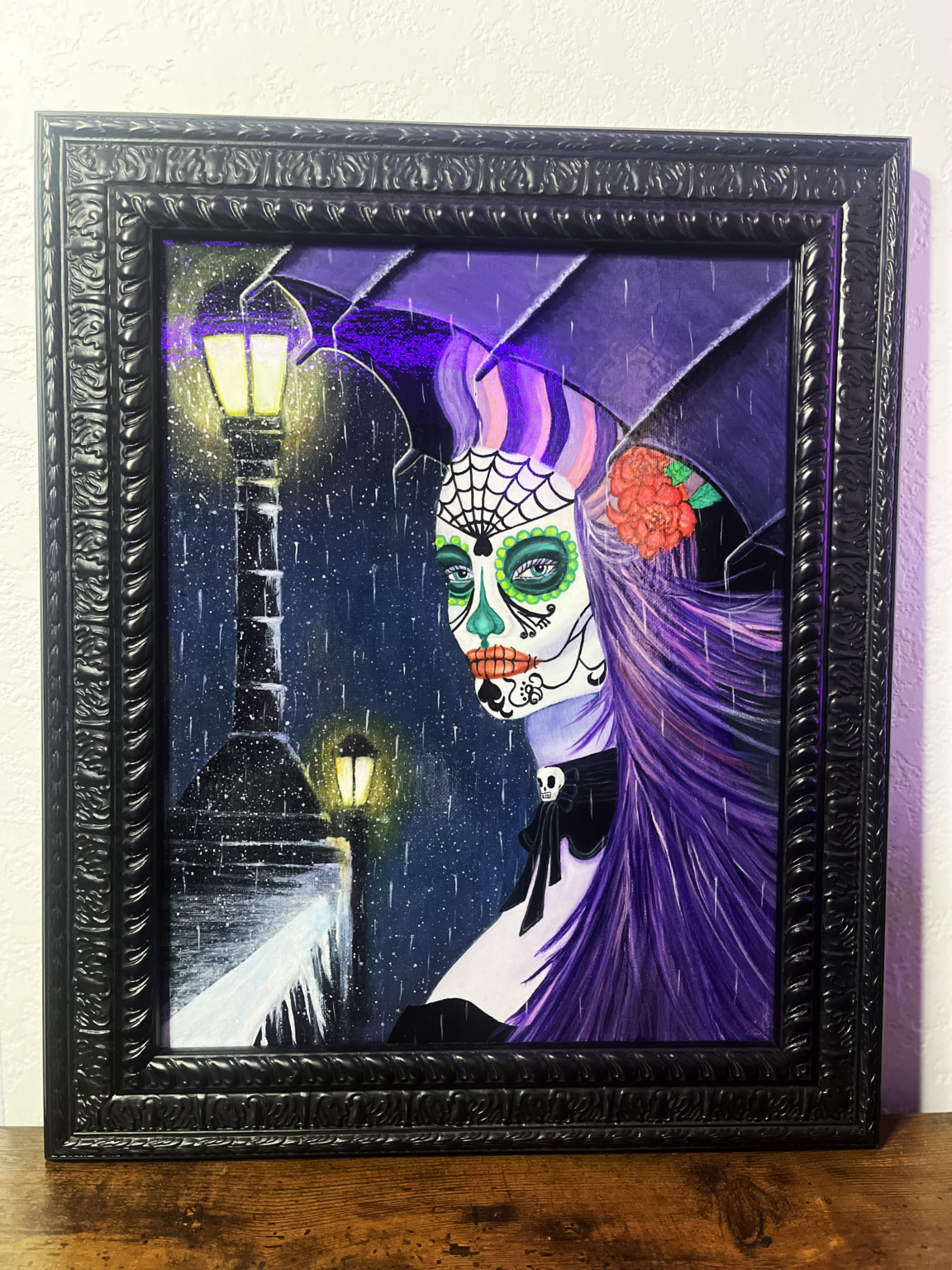"Night of the Beauty" an original painting by The Dreaming Siren, depicting a woman with a Día de los Muertos face painting, vibrant purple hair, and a red flower, set against a nighttime street scene with rain and street lamps.